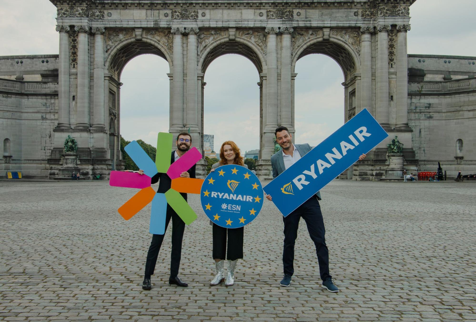 Three people, standing in front of a large stone arch monument, each holding oversized colourful props: the Erasmus Student Network logo, a circular Ryanair–ESN logo, and a rectangular Ryanair sign.